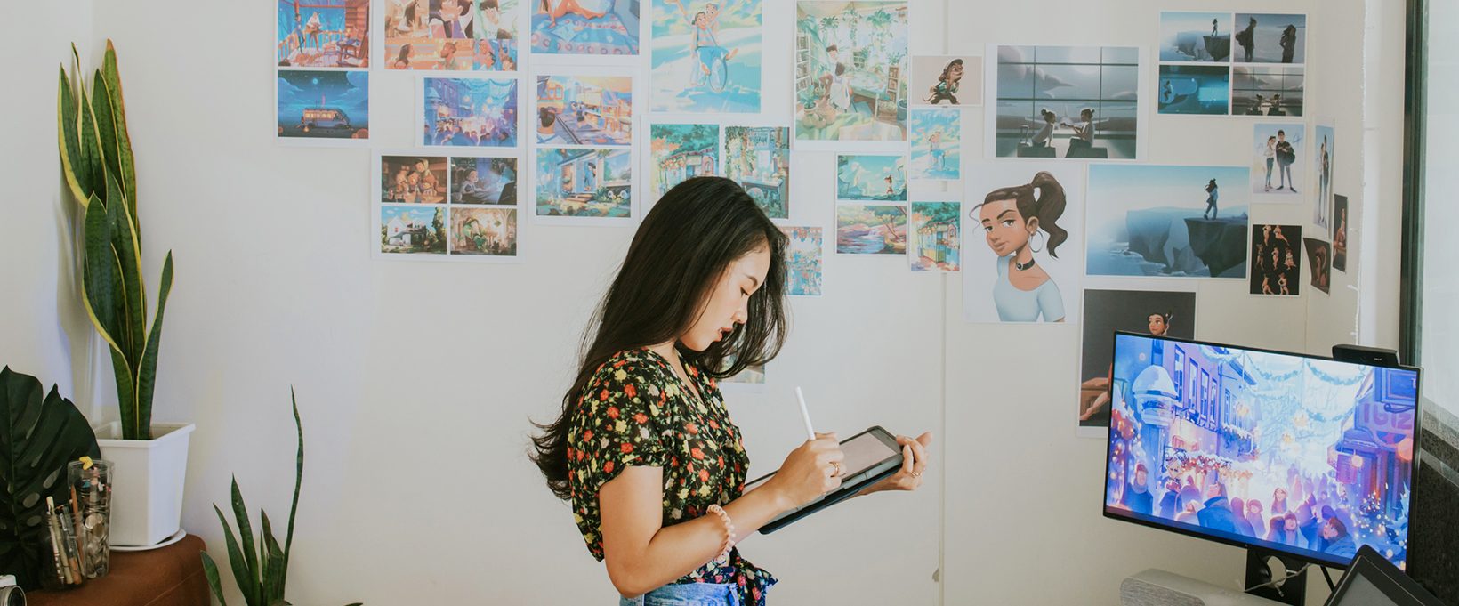 A designer is using a digital drawing pad in a design studio with many sketches stick on the wall next to her.