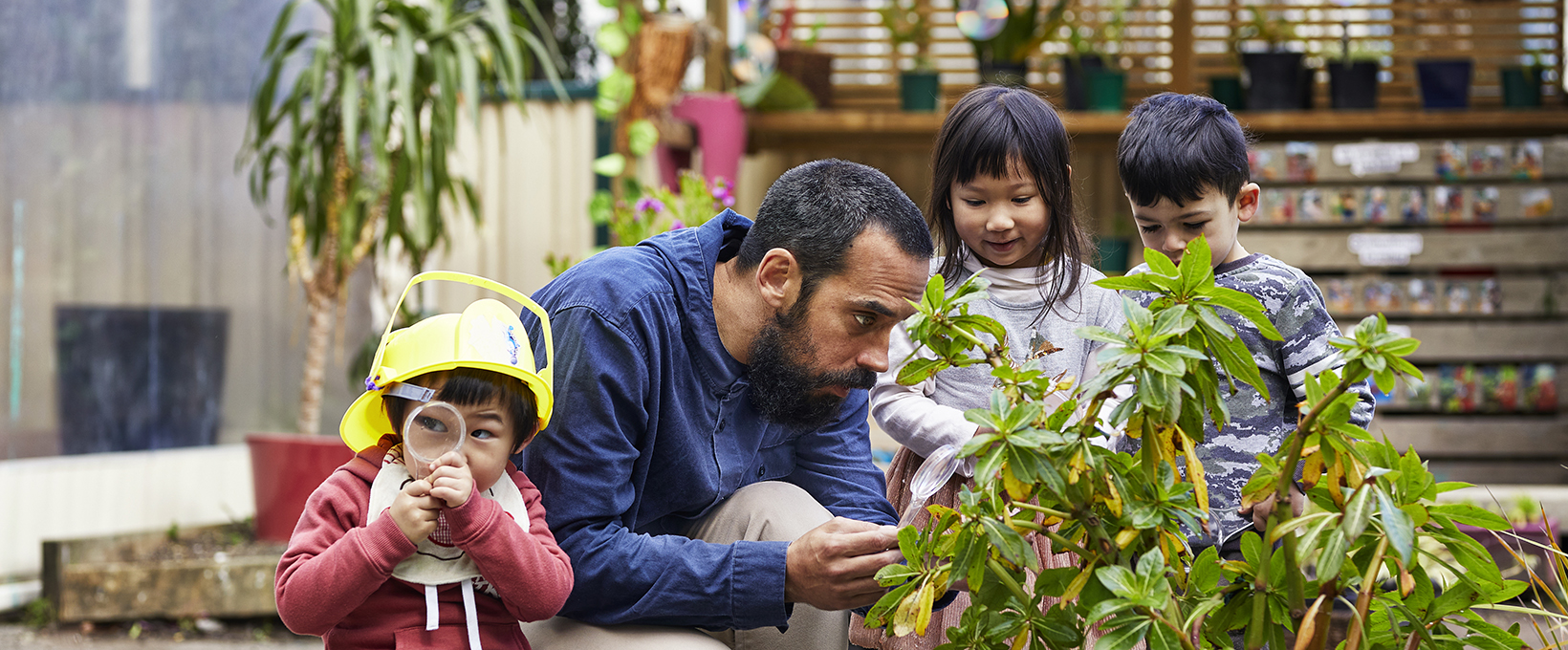 An early childhood centre teacher is inspecting a plant with three chidren (tamariki) in an outdoor environment 