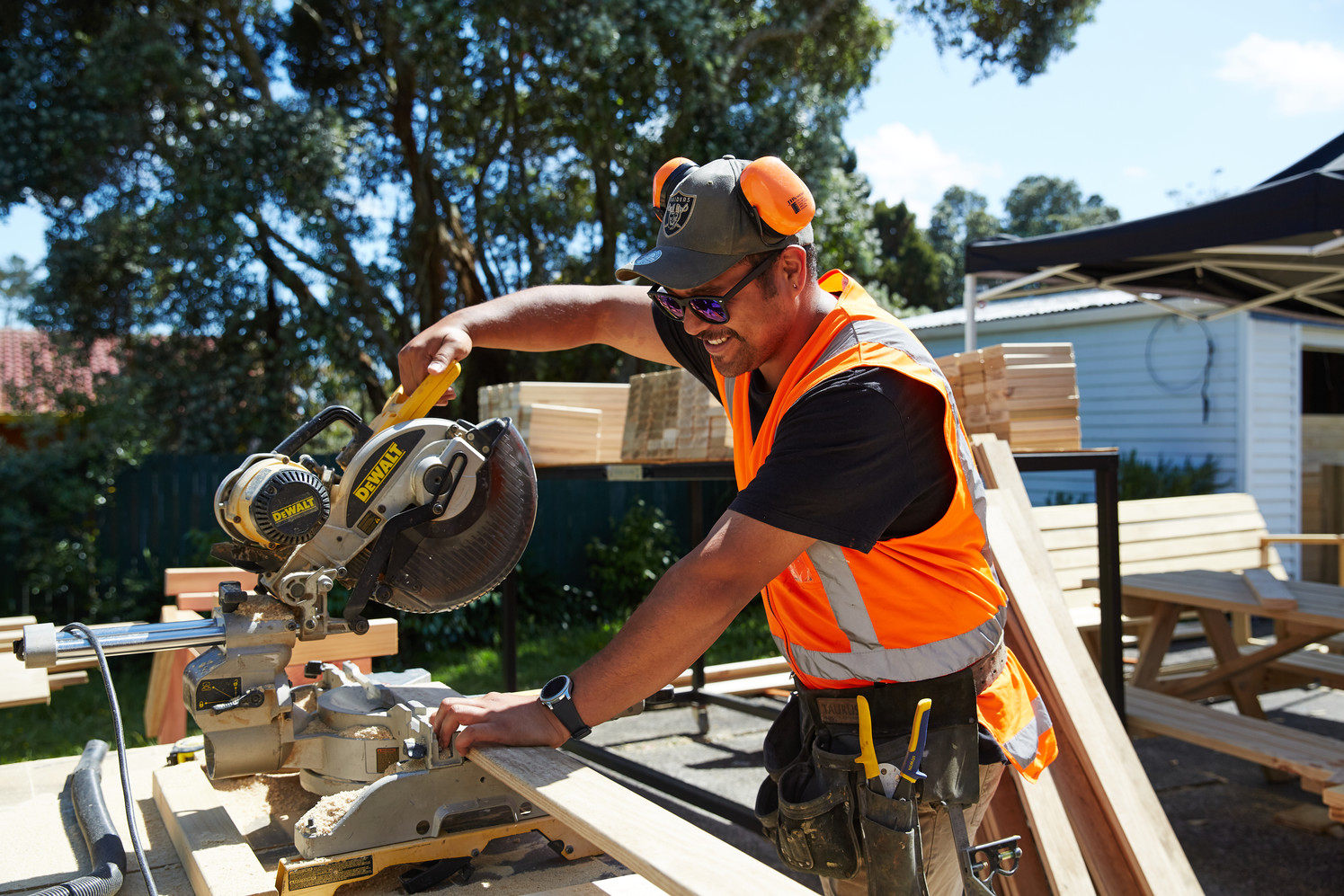 Unitec Building and Carpentry student in orange hi-vis gear using a saw to cut a plank of wood