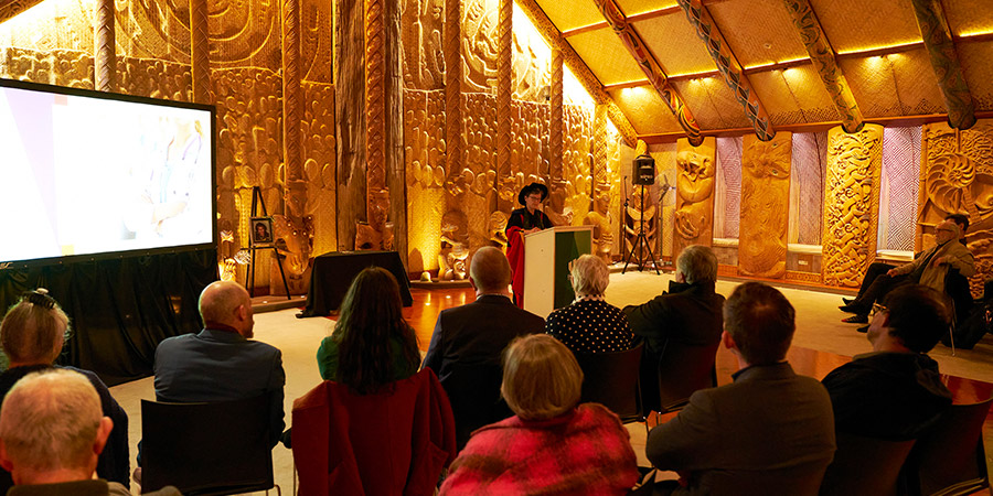A wide angle shot showing Professor Helen Gremillion presenting her Professorial Address on interdisciplinary research to a captivated audience inside the wharenui at Te Noho Kotahitanga marae
