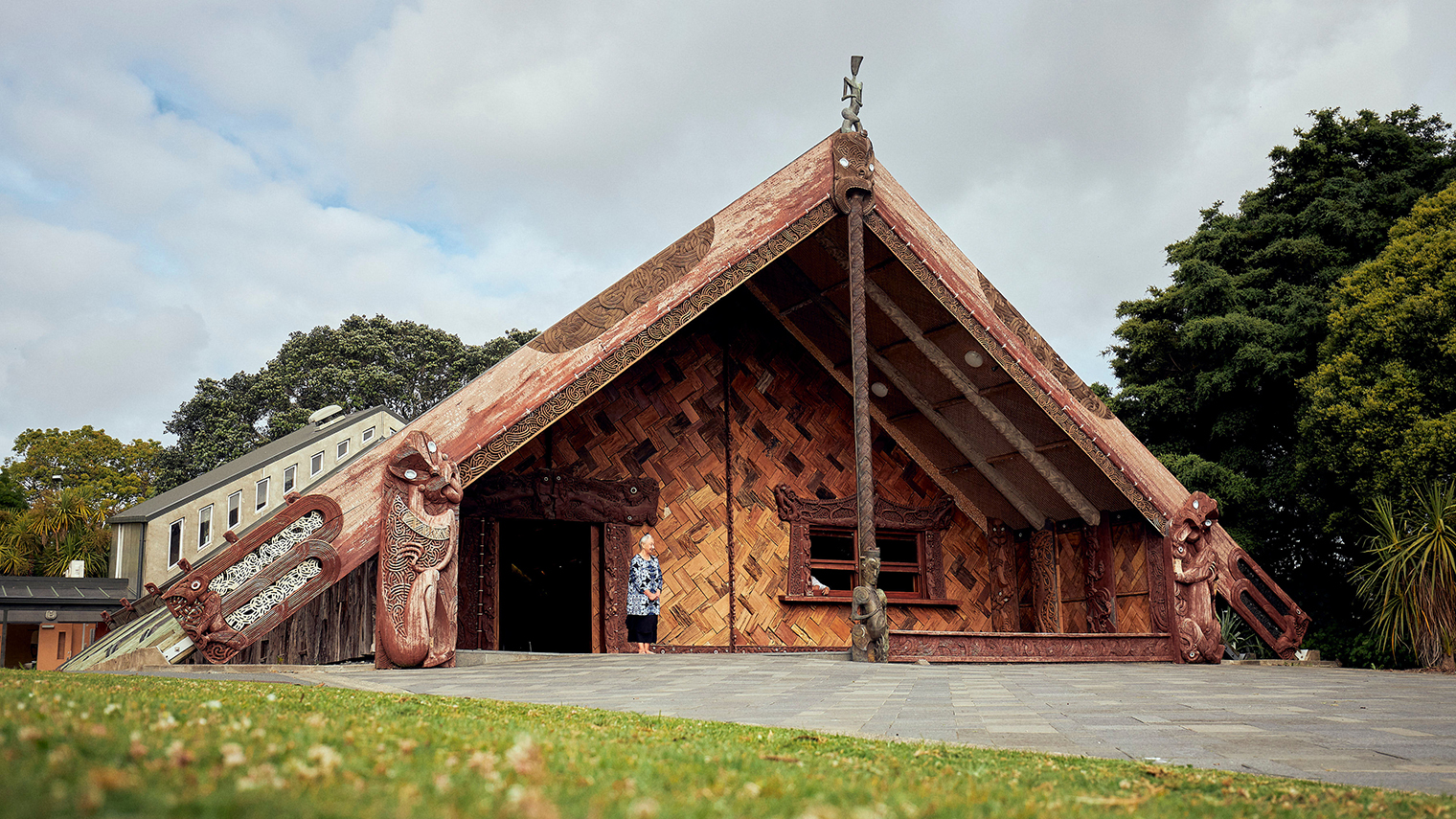 Unitec Te Noho Kotahitanga Marae with intricately carved wooden exterior, steep pitched roof, and surrounding greenery, under a partly cloudy sky.