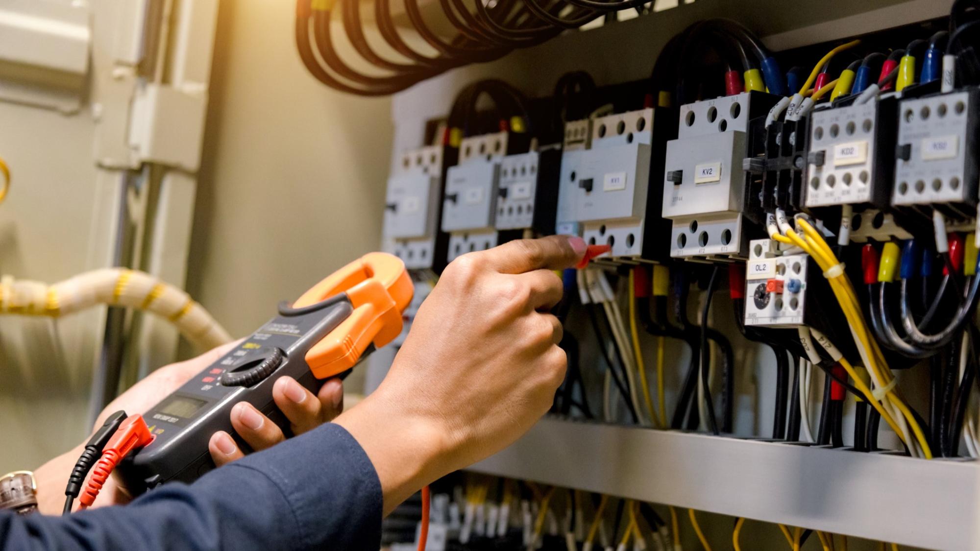 A person using a clamp meter to test electrical components in a power distribution panel filled with wires and circuit breakers.