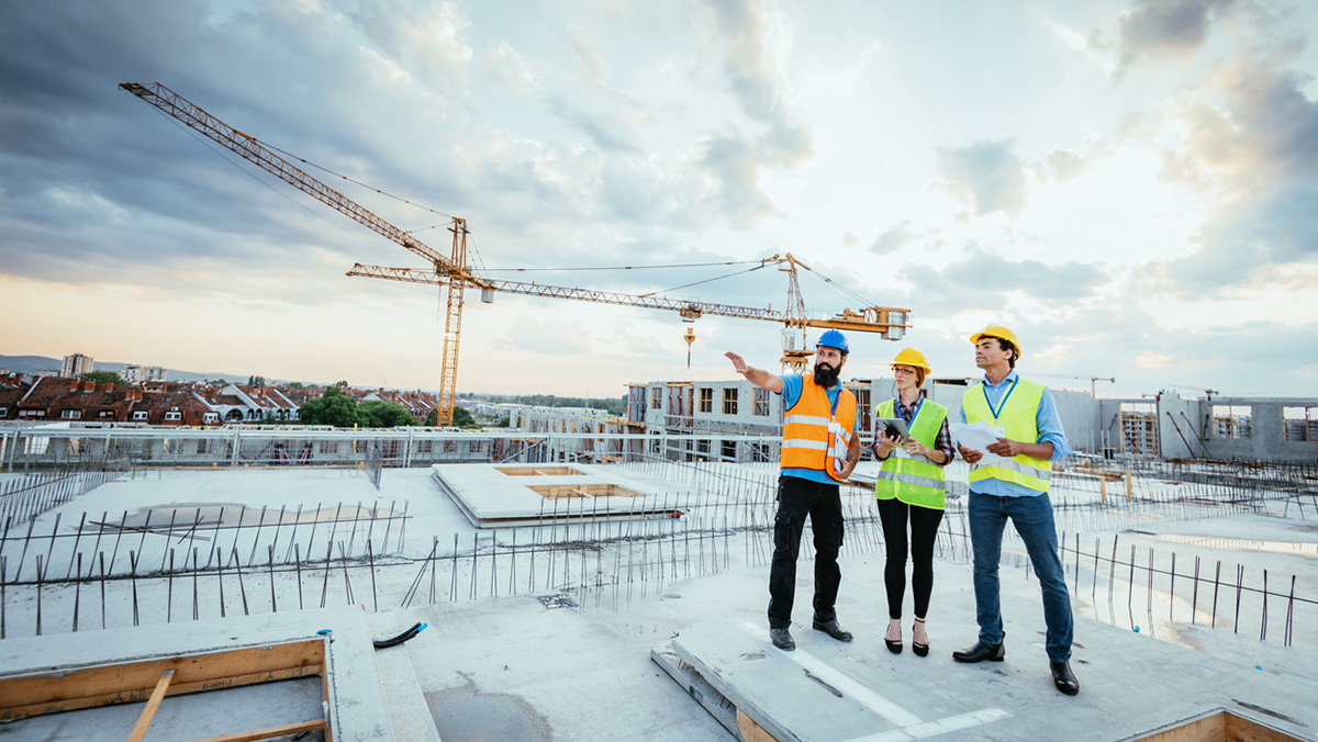 Three construction workers in high-vis vests and hard hats stand on a busy site, one pointing toward cranes and unfinished buildings under a partly cloudy sky.