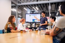 Group of people at table with presenter standing explaining information on large screen