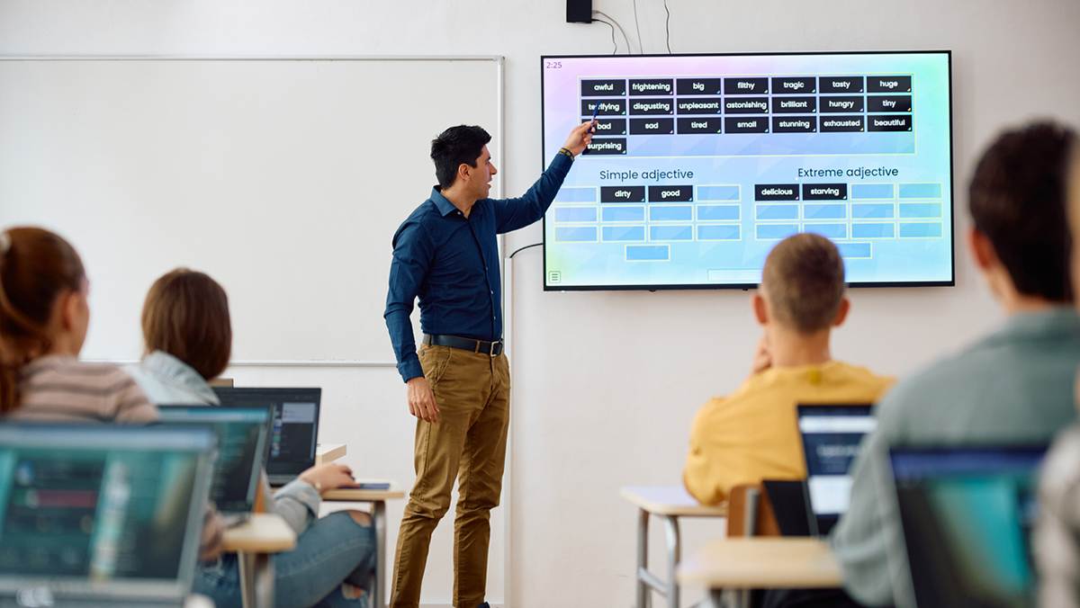 Educator uses AI-generated vocabulary list on a projector while students engage with laptops in a modern classroom, showcasing GenAI-enhanced learning.