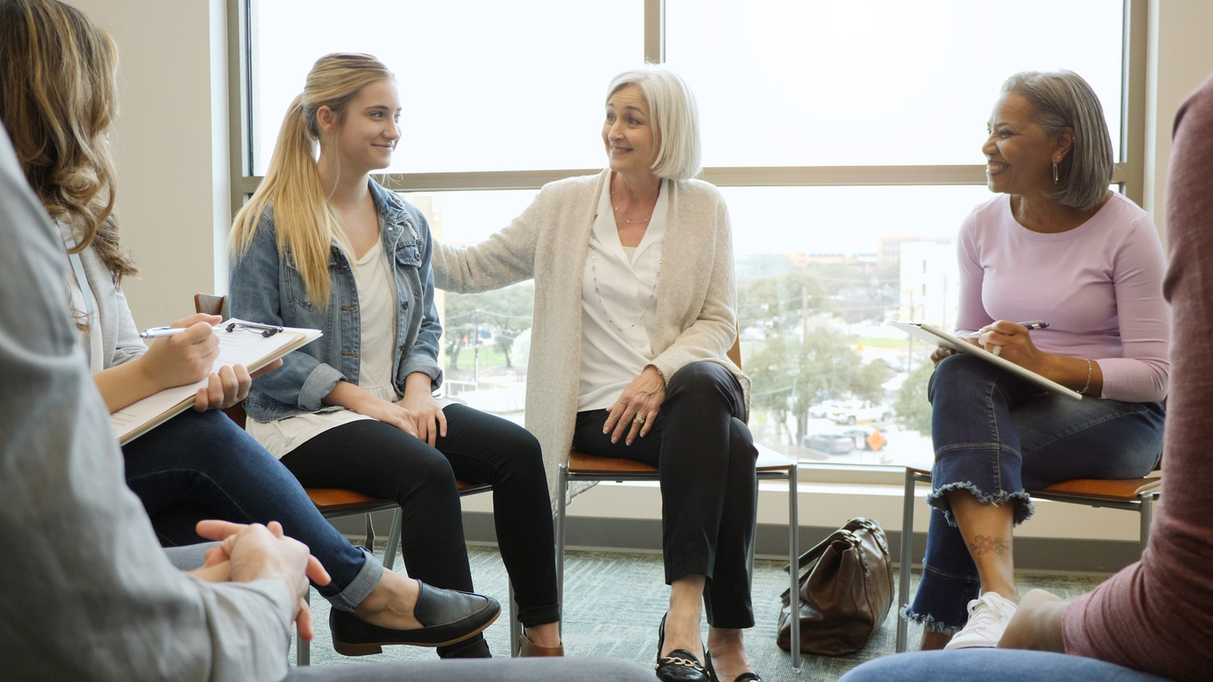 Group of diverse women seated in a circle for a supportive conversation or therapy session near a large urban-view window, displaying attentive and empathetic engagement.