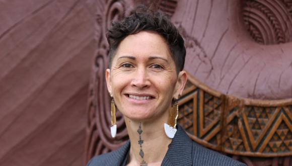 Portrait of Deb Heke smiiing while standing in front of Ngākau Māhake, the whare nui at Te Noho Kotahitanga marae