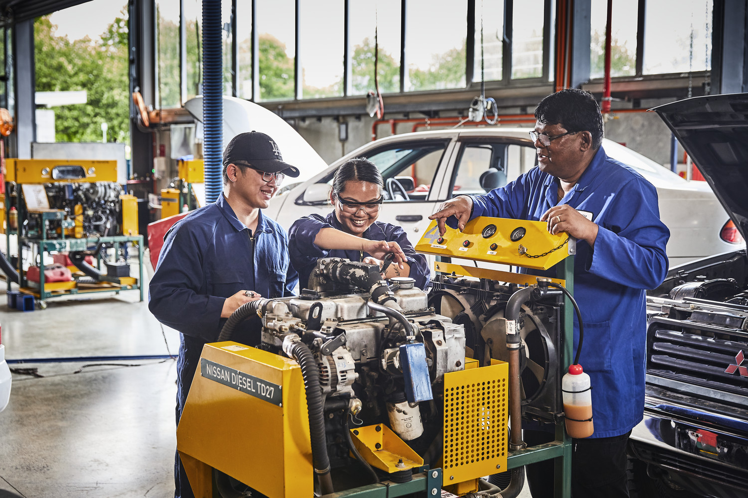 3 automotive engineering students at Unitec working on a car engine together with a workshop classroom in the background