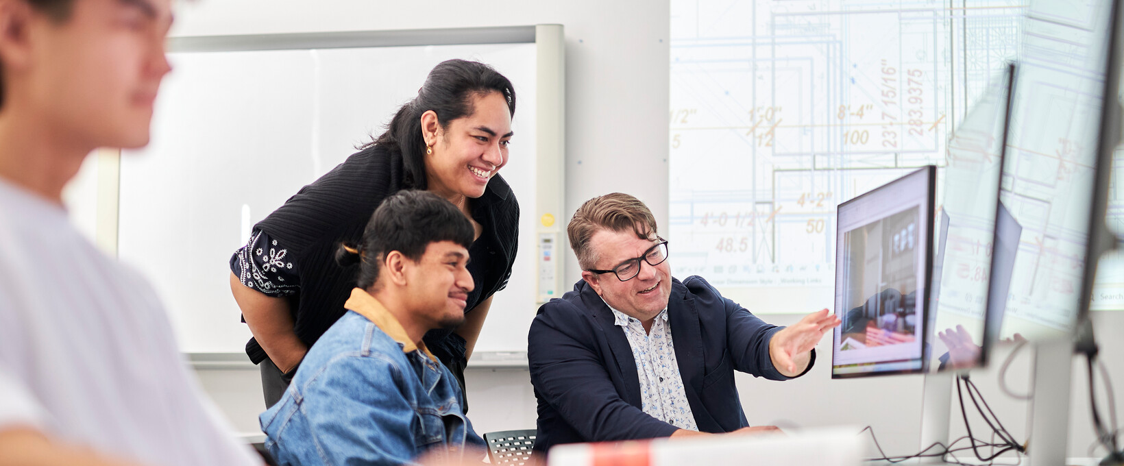 A tutor and students collaborate around a computer, reviewing architectural drawings and digital designs in a classroom setting.