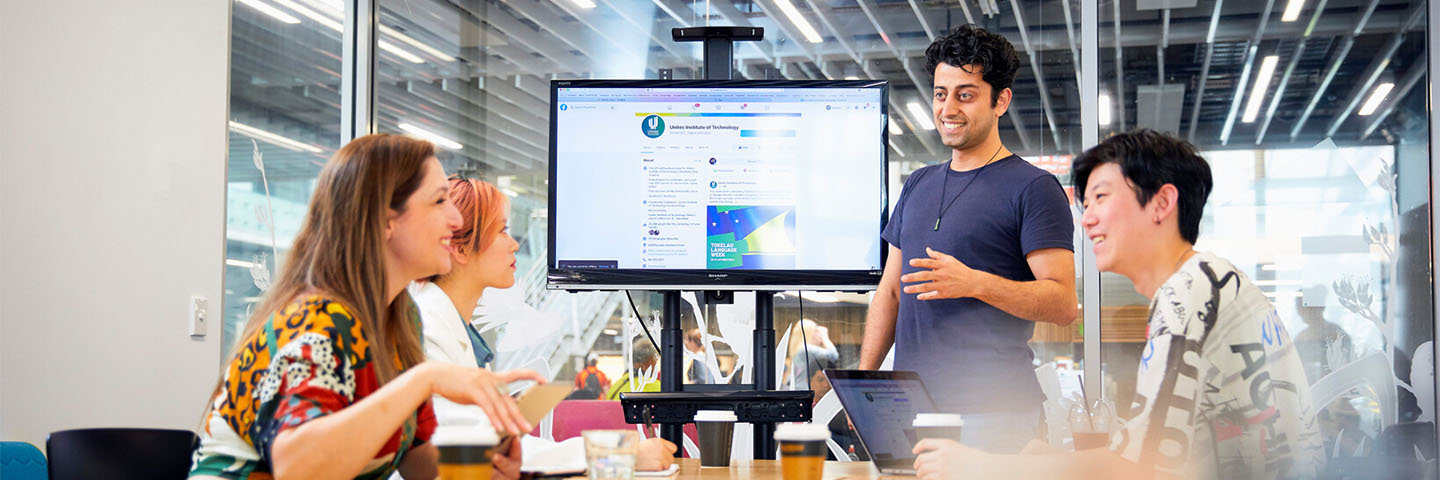 A group of students sit at a meeting table while another student gives a presentation.