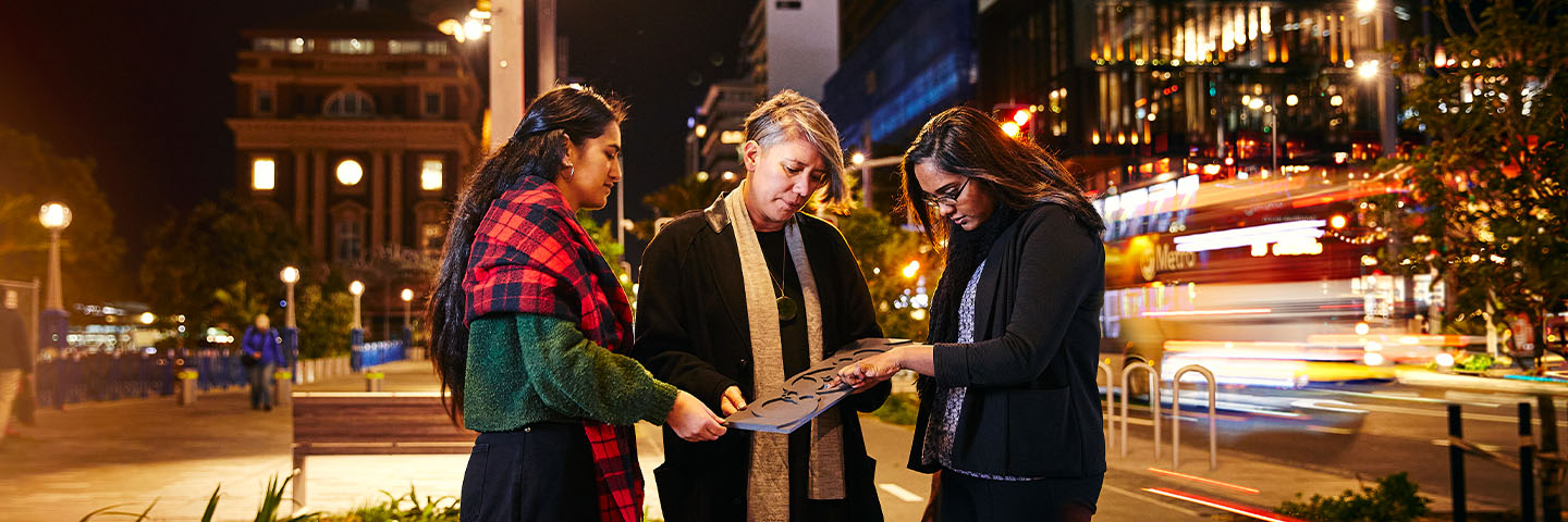 Three students examine some plans on a busy nighttime street.