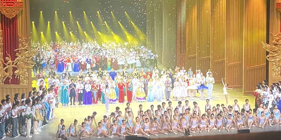 Hundreds of dancers in traditional Chinese dance attire assemble on stage in a theatre at the end of a performance.