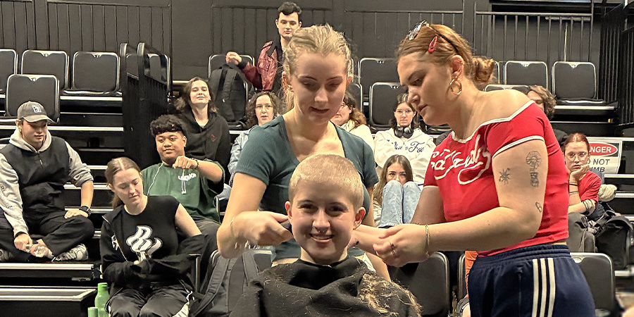 Brooke Wilson in the middle of having her head shaved by two fellow students, in front of an audience of friends, kaimahi and classmates in a black box theatre.