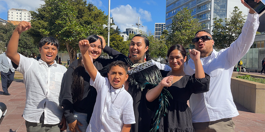 Corren Awheto in graduation attire and a korowai, surrounded by her family, raising their fists triumphantly in the air and smiling.