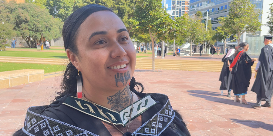 Corren Awheto in black graduation attire decorated with a traditional Māori pattern in white, smiling outdoors with other graduates in the background.