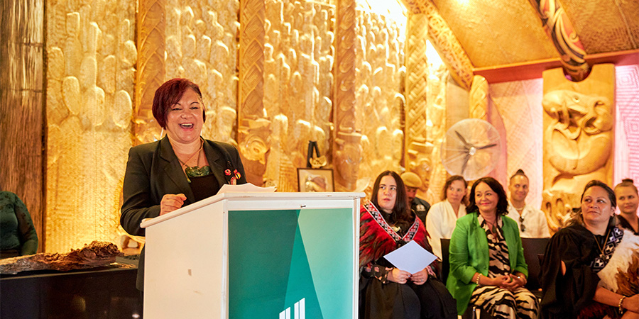 Tonina Ngatai stands behind a lectern in the meeting house on Te Noho Kotahitanga Marae, giving a speech to an audience. 