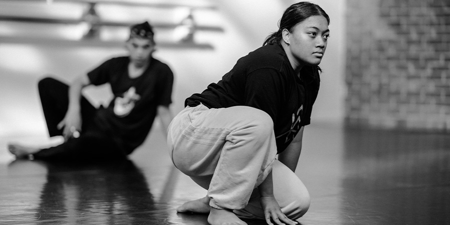 A black and white image of Praise Tupa'i crouching during a dance rehearsal in front of another dance student.