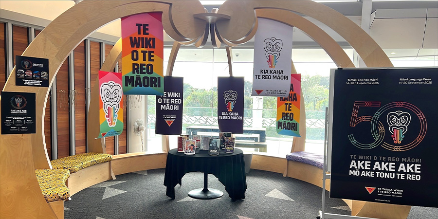 A colourful display of posters, leaflets and books advertising Te Wiki o Te Reo Māori in a sunny area of the Te Puna student centre with pillars carved with koru at the top that meet in the middle.