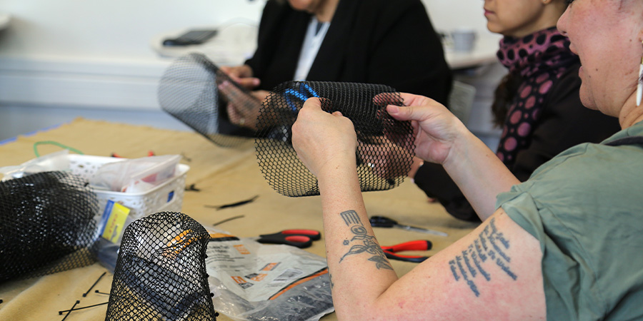 People sit round a table during a craft workshop, with basket-making materials, pliers and wire cutters on the table. A woman in the foreground with tattoos on her arms holds some of the basket mesh. 
