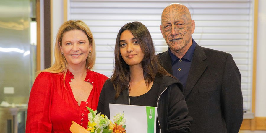 Dr Roseanne Ellis and Kaihaūtu, Harawira Pearless stand on either side of winner Maria Gomes, who is holding flowers and a certificate.