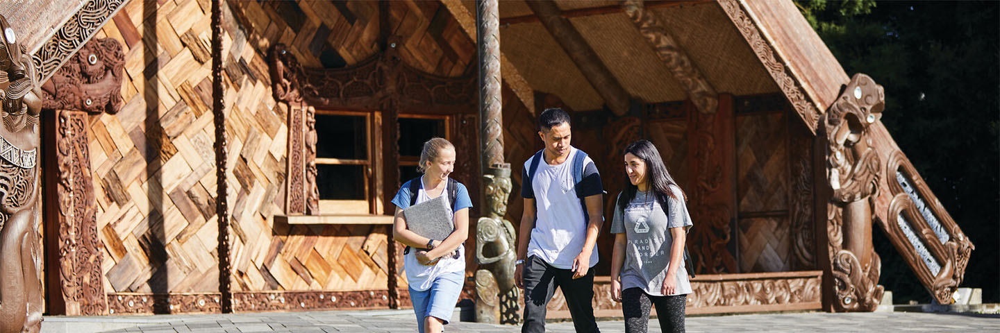 Three students walk and talk in the sunshine outside the Unitec Marae meeting house.