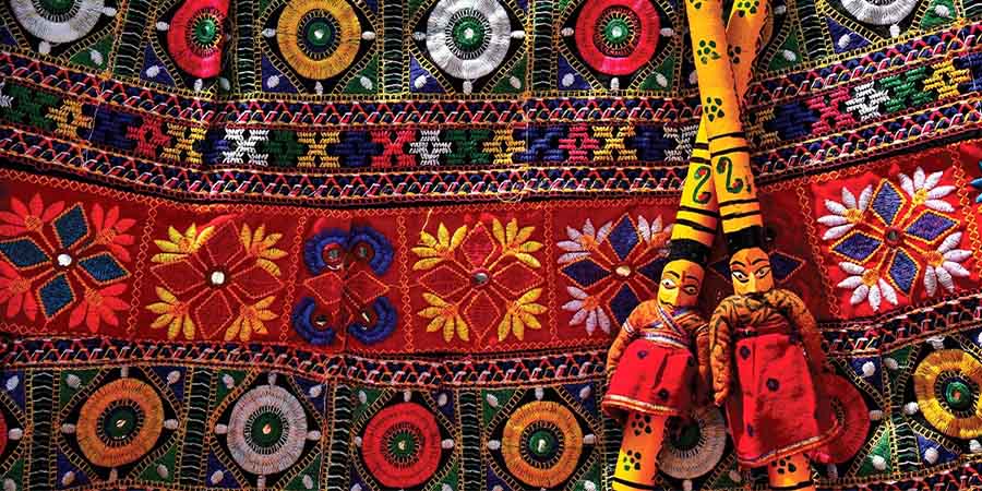 Two dolls on decorated wooden poles lie on top of an intricately embroidered cloth. 