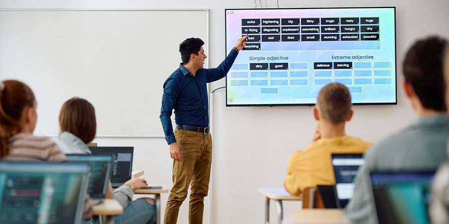 A teacher stands at the front of a class, pointing to an interactive whiteboard.