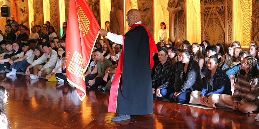 Harawira Pearless waves a school flag in front of assembled students on the floor, in te wharenui o Te Noho Kotahitanga Marae.