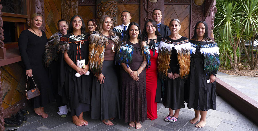 Māori graduates in graduation attire and korowai pose together outside te wharenui.