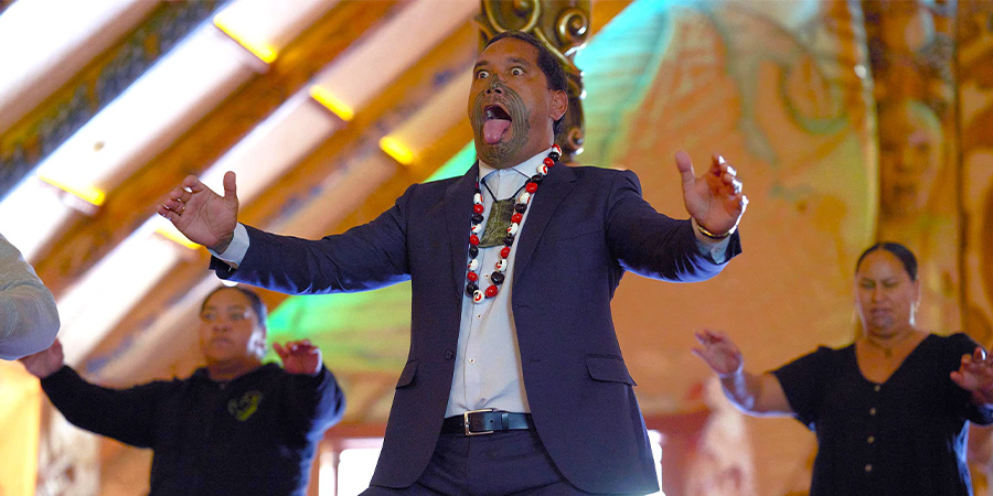 A man with ta moko on his face and two other people perform a haka inside te wharenui.