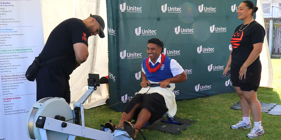 A student in Polynesian attire uses a rowing machine watched by a staff member in sportwear. 