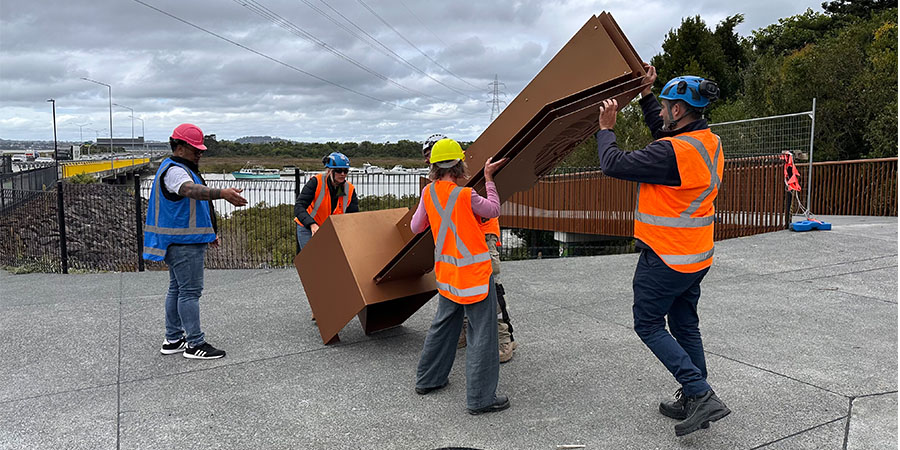 A team of people in high-vis vests and hard hats work together to erect the large metal sculptural marker.