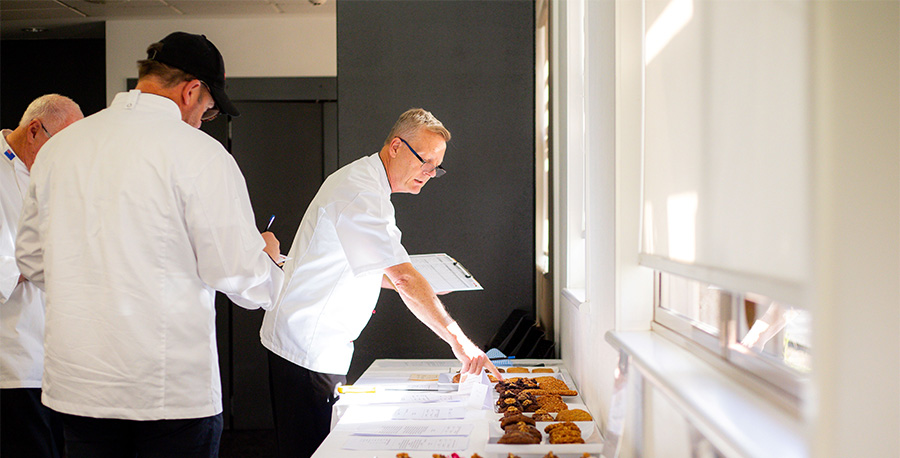 Judges in chef's uniform examine entries in the cooking competition on a long table.