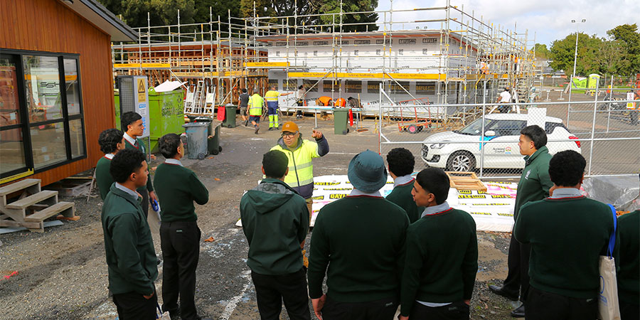 A group of students in school uniform stand at the edge of Unitec's trades building site listening to a teacher.