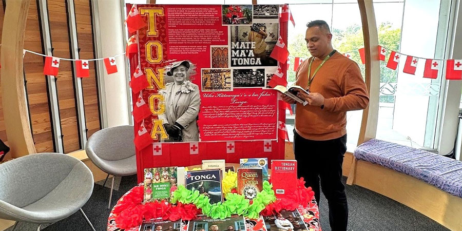 A man reads a book in front of a display about Tongan language and culture, with a table full of books about Tonga in front of him.