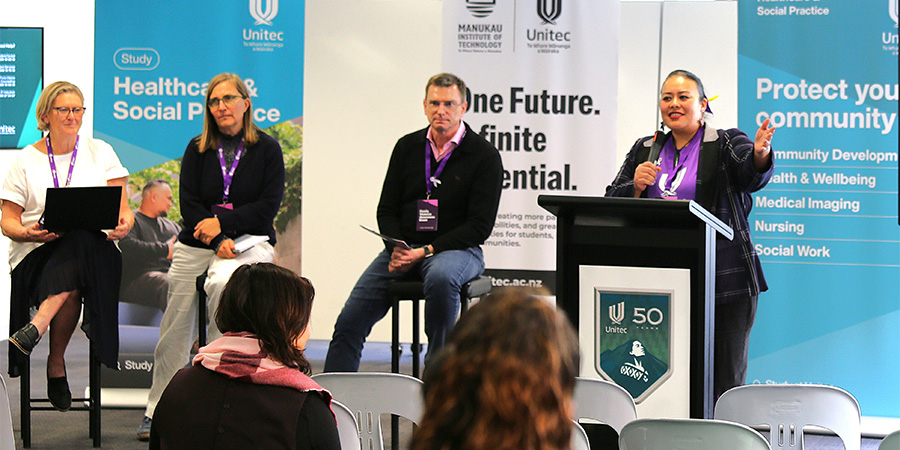 A woman in a suit stands at a lectern introducing three people seated on tall stools to an audience.