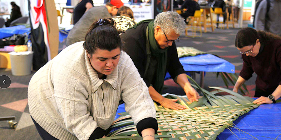 Three people weave tukutuku together.