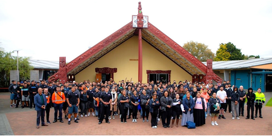 A large group of people smile for the camera outside the wharenui on Ōtara campus.