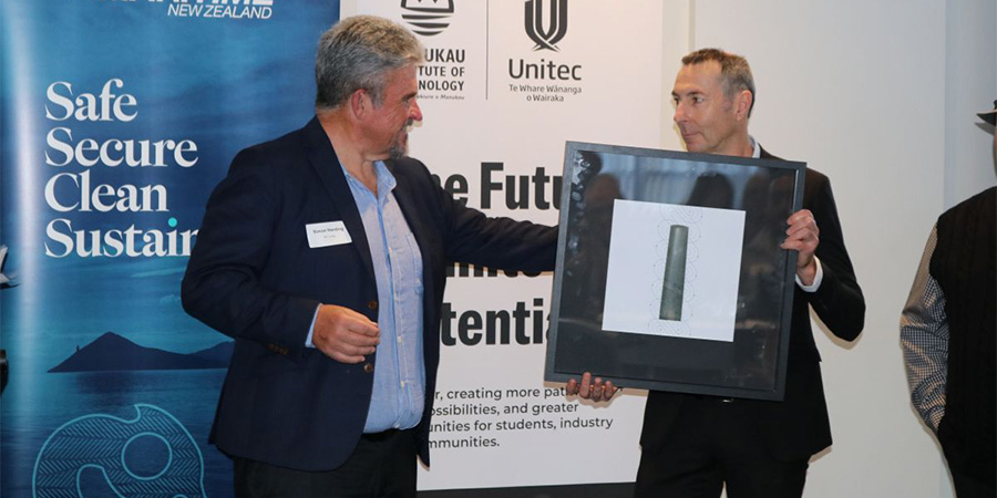 Two men in suits during a presentation, one holding a framed taonga.