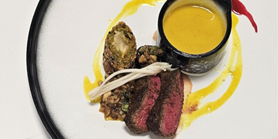 A beautifully presented plate of food including seared beef and a bowl of sauce.