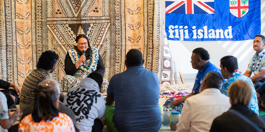 A group of people in traditional Fijian attire sit in a circle listening to a woman speak.
