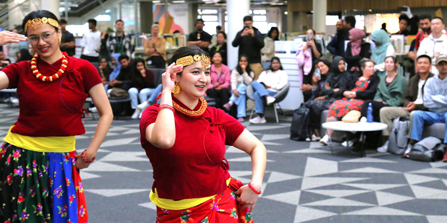 Two women in traditional Nepali attire dance in front of a large audience in a library space.