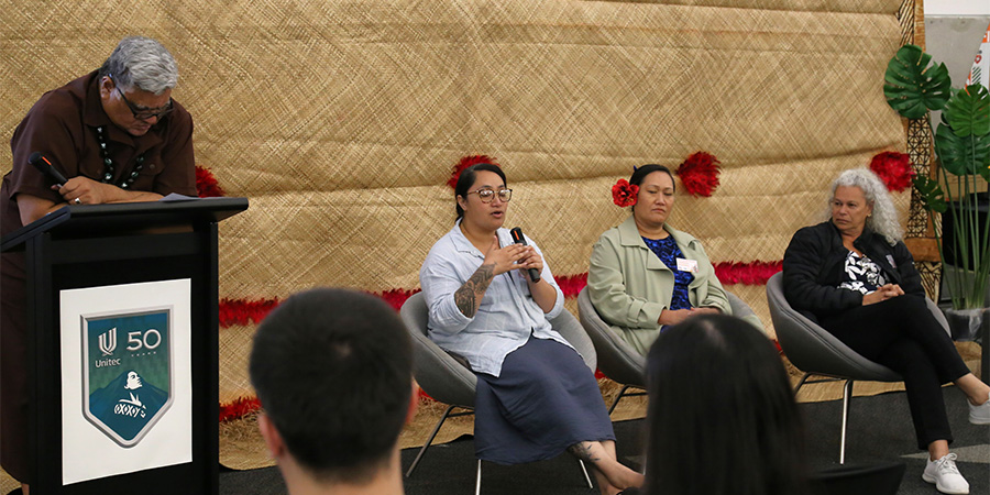A man stands at a lectern and three women sit on chairs beside him, while one of the women speaks into a microphone.
