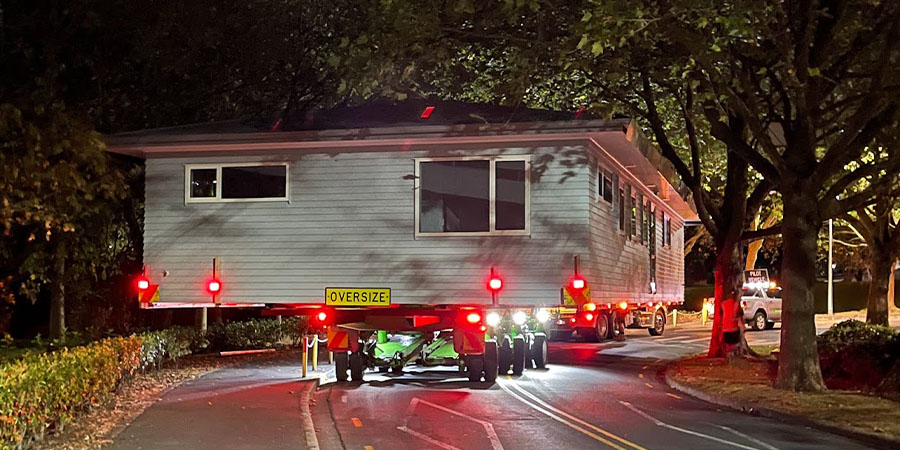 A house with a sign on the back saying 'oversize' loaded on a truck, being driven out of Unitec's Mt Albert campus gate 4 at night, with multiple lights.